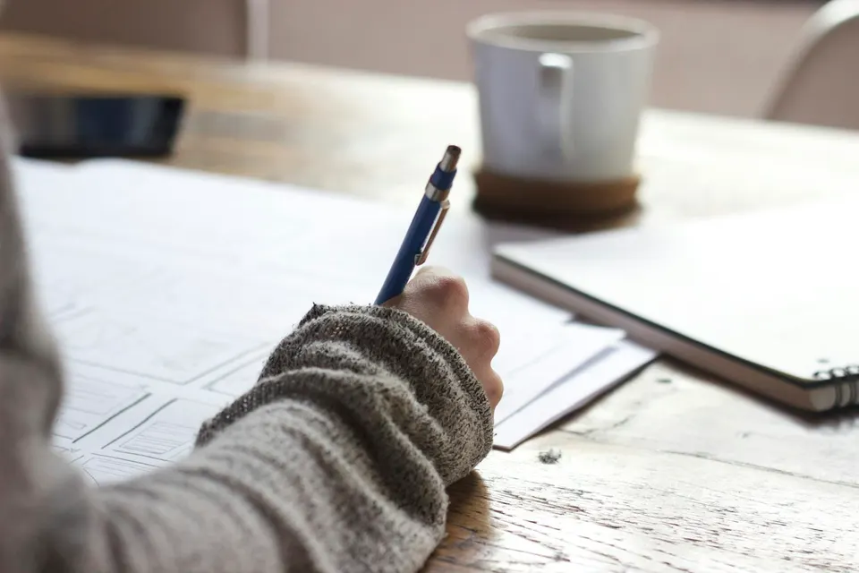 A person writing notes beside a laptop and a cup